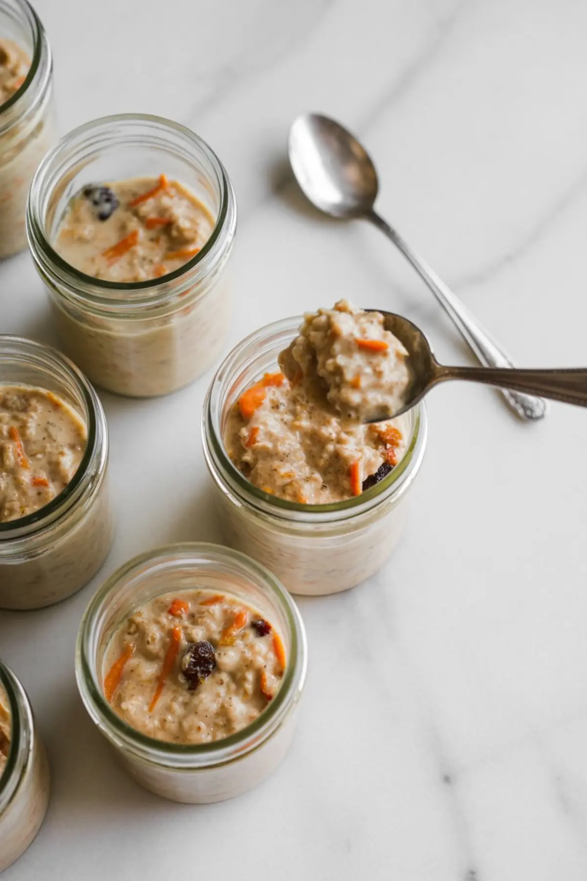 Glass jars filled with creamy carrot cake overnight oats, showing visible carrot shreds and raisins, with one spoonful lifted above a jar on a marble surface.

