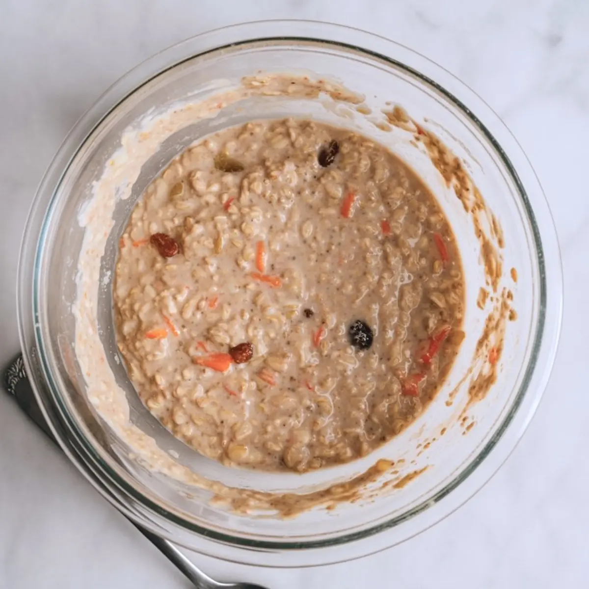 Overhead view of a mixing bowl filled with blended carrot cake overnight oats, showing visible oats, raisins, and shredded carrots in a creamy mixture.
