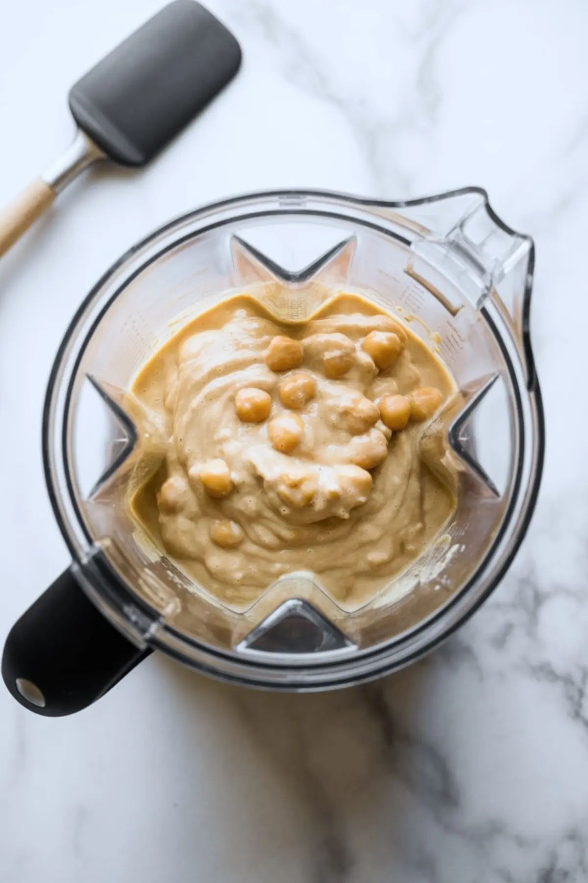 Thick chickpea muffin batter with whole chickpeas visible inside a blender, placed on a white marble counter with a silicone spatula nearby, ready for blending.