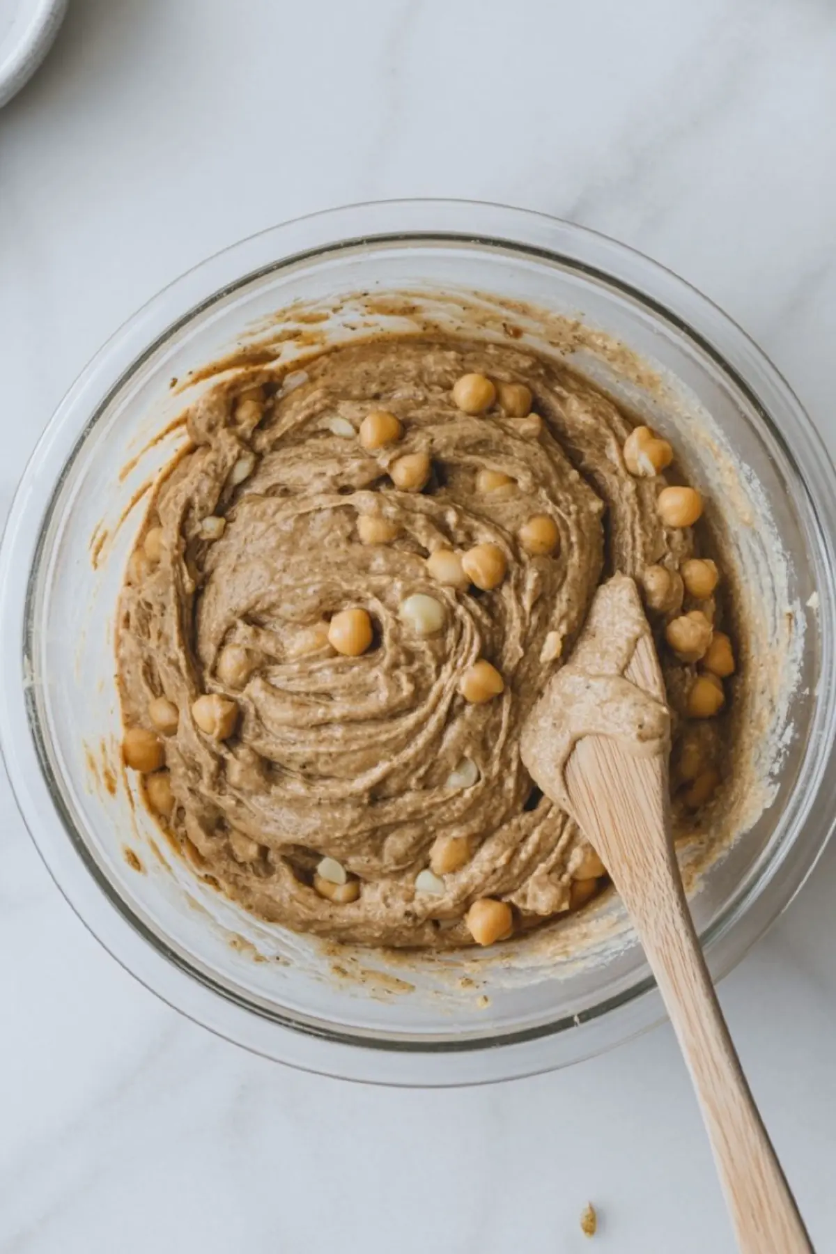 Top-down view of thick chickpea muffin batter in a glass mixing bowl, with visible whole chickpeas and a wooden spatula resting in the mix, ready to be scooped.