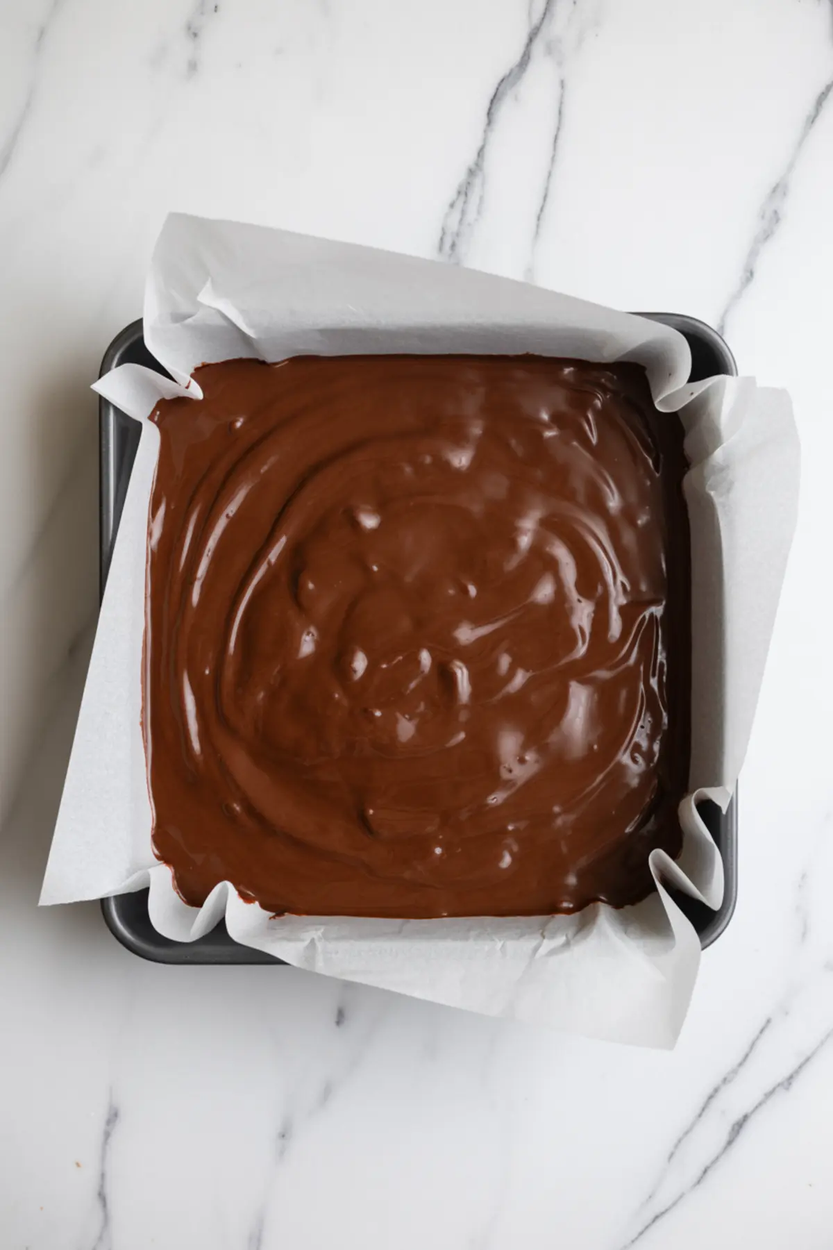 Uncut pan of chocolate coconut fudge in a square baking tray lined with parchment paper. The glossy melted chocolate top has smooth ripples, indicating a freshly poured layer. Shot captures the prep stage for homemade low-sugar fudge bars.
