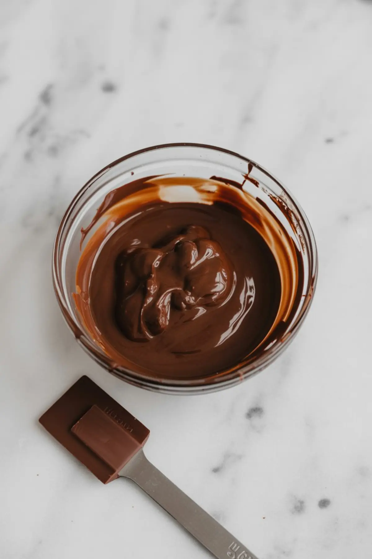 Glass bowl of melted dark chocolate on a marble surface, with a silicone spatula coated in chocolate beside it. The glossy, smooth texture is ready for layering in low-carb dessert recipes or keto fudge prep.
