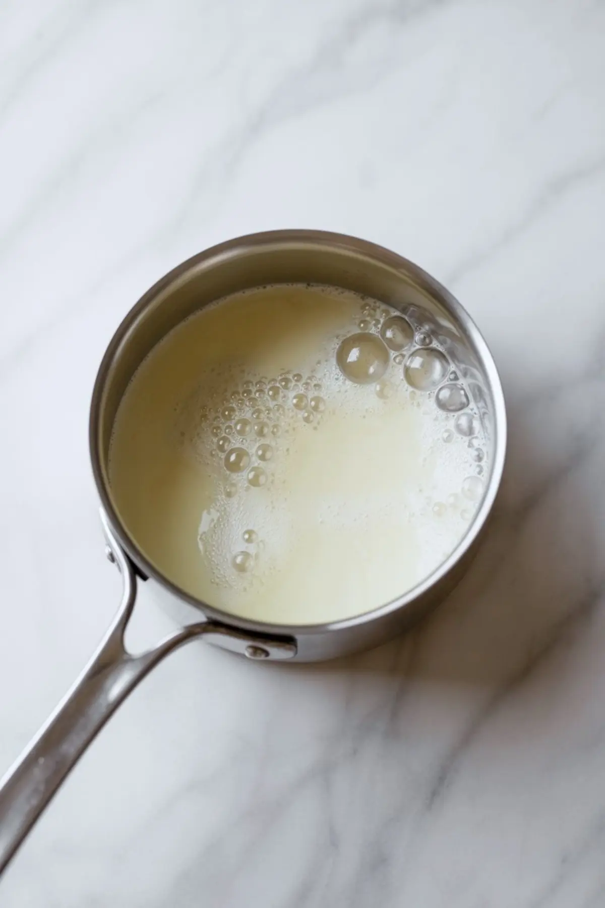 A stainless steel saucepan filled with hot cream shows visible steam bubbles forming on the surface, resting on a white marble countertop.