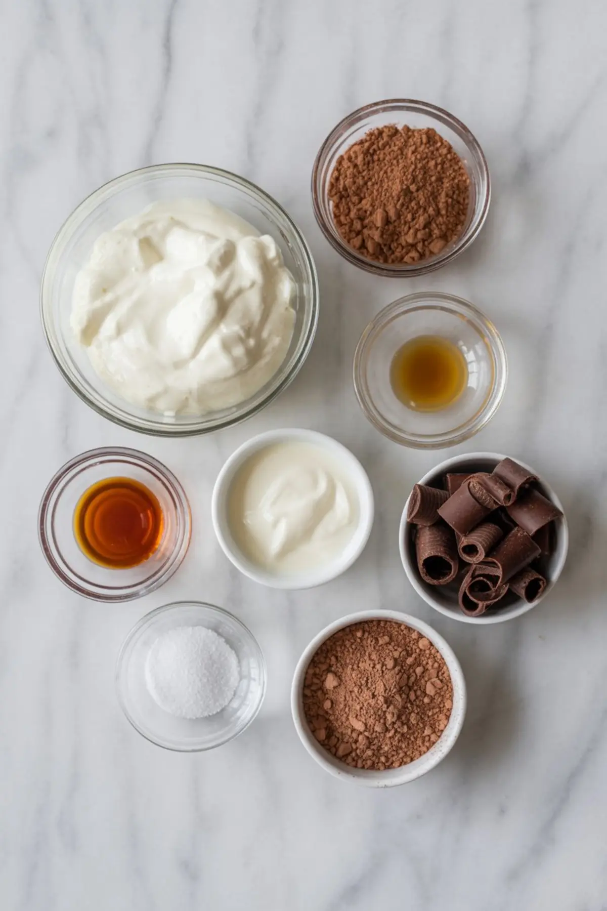Overhead view of chocolate Greek yogurt ingredients on a marble background, including Greek yogurt, cocoa powder, vanilla extract, chocolate curls, sweetener, and salt in small glass and ceramic bowls.