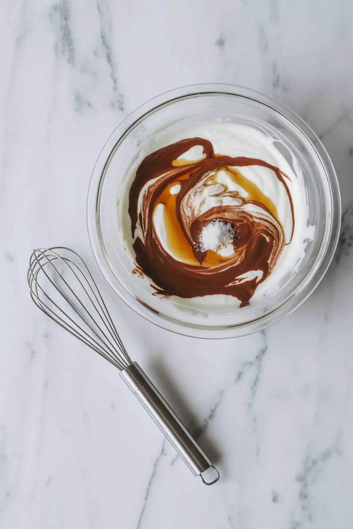 Top-down view of a mixing bowl filled with Greek yogurt, cocoa powder, sweetener, vanilla, and salt before being whisked together, with a metal whisk placed beside the bowl on a marble surface.