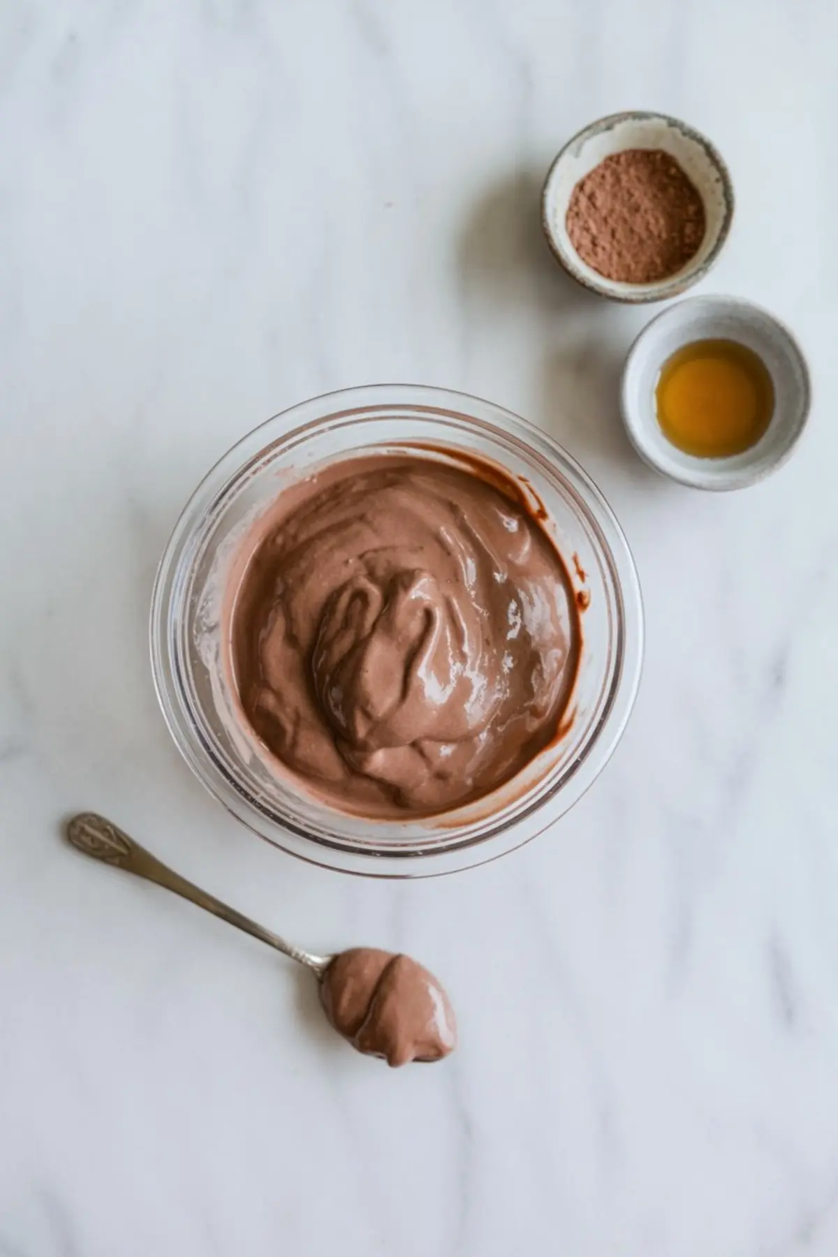 Creamy chocolate Greek yogurt blended in a glass bowl with a small spoonful beside it, surrounded by bowls of cocoa powder and vanilla extract on a light marble countertop.