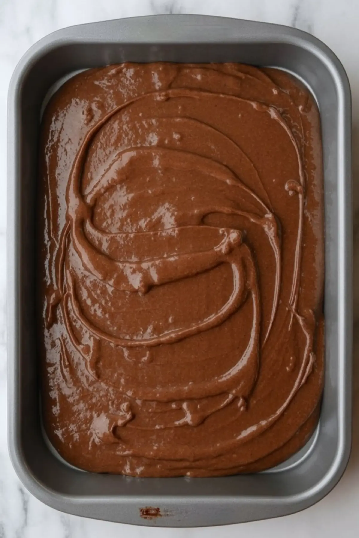 Rectangular baking pan filled with unbaked chocolate cake batter, evenly spread across the surface with visible swirls and a glossy texture.