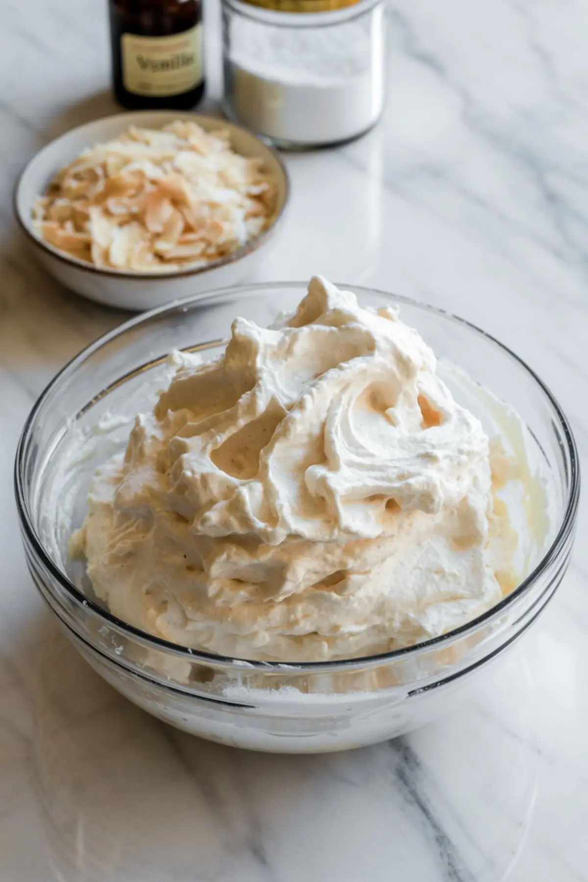 Glass bowl filled with fluffy coconut whipped frosting on a marble counter, surrounded by ingredients like toasted coconut flakes, powdered sugar, and vanilla extract in the background.
