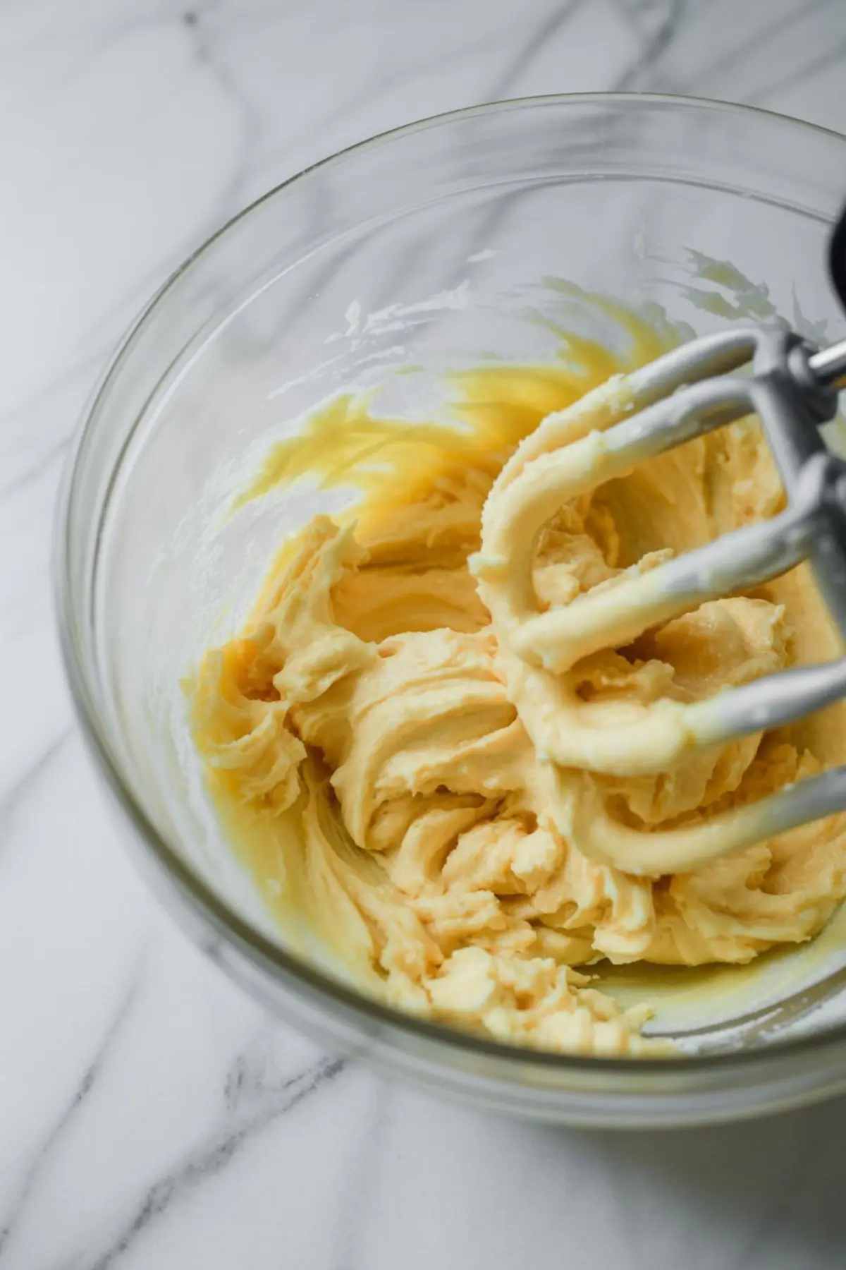 Creamed butter mixture in a glass bowl being whipped with metal beaters, captured on a white marble countertop during cake preparation.
