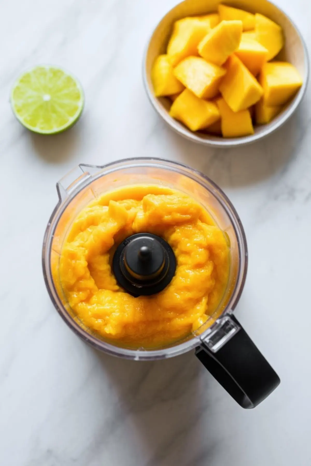 Food processor with smooth mango puree beside a bowl of cubed mango and a halved lime on a marble surface.