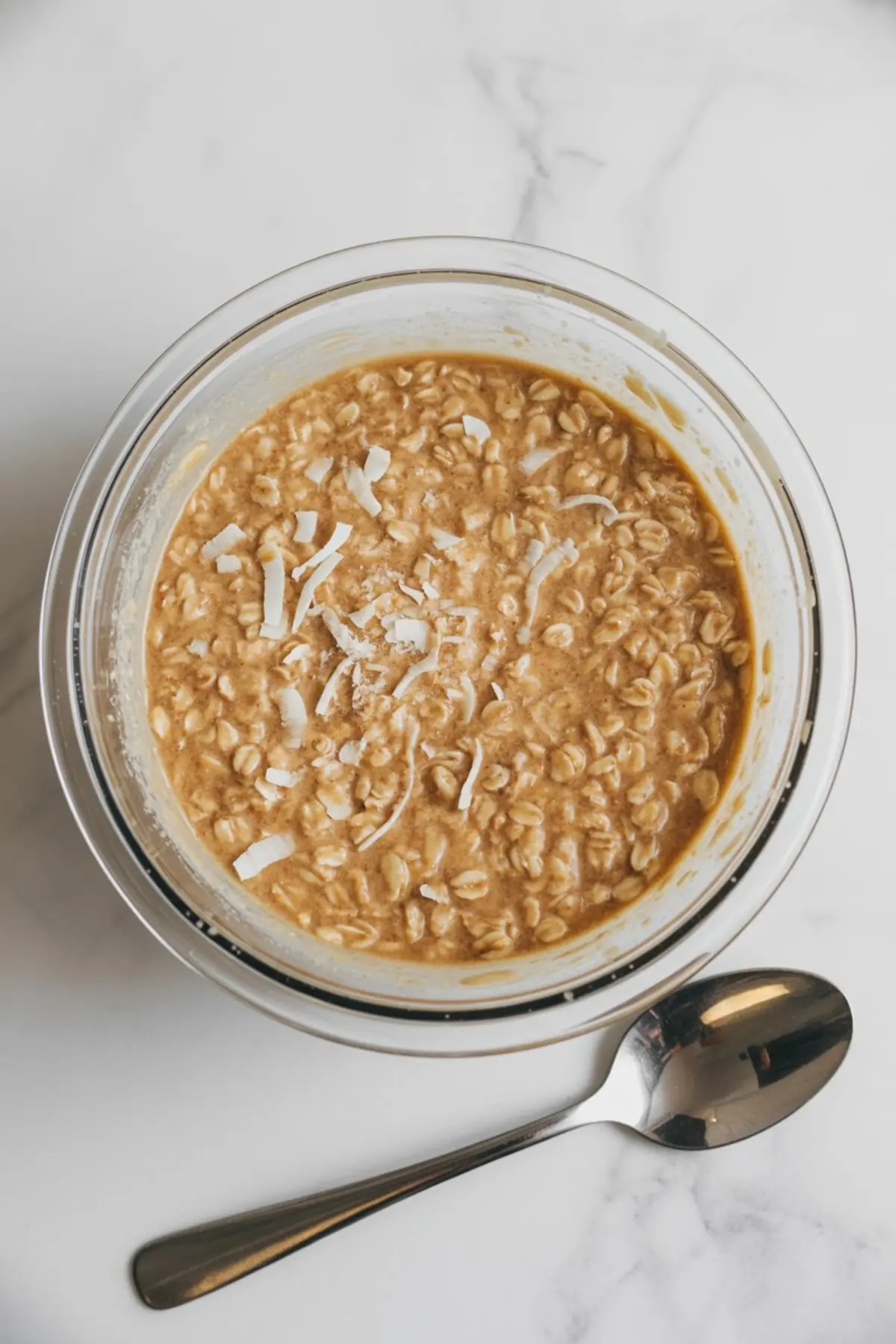 Top view of a glass bowl filled with a mixture of oats and shredded coconut in a creamy base, set on a marble countertop. A stainless steel spoon lies beside the bowl, indicating preparation for coconut-flavored overnight oats.
