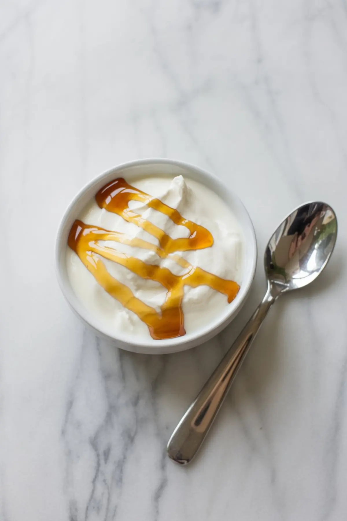 Small white bowl filled with thick Greek yogurt topped with a drizzle of maple syrup, placed on a white marble surface next to a reflective metal spoon.
