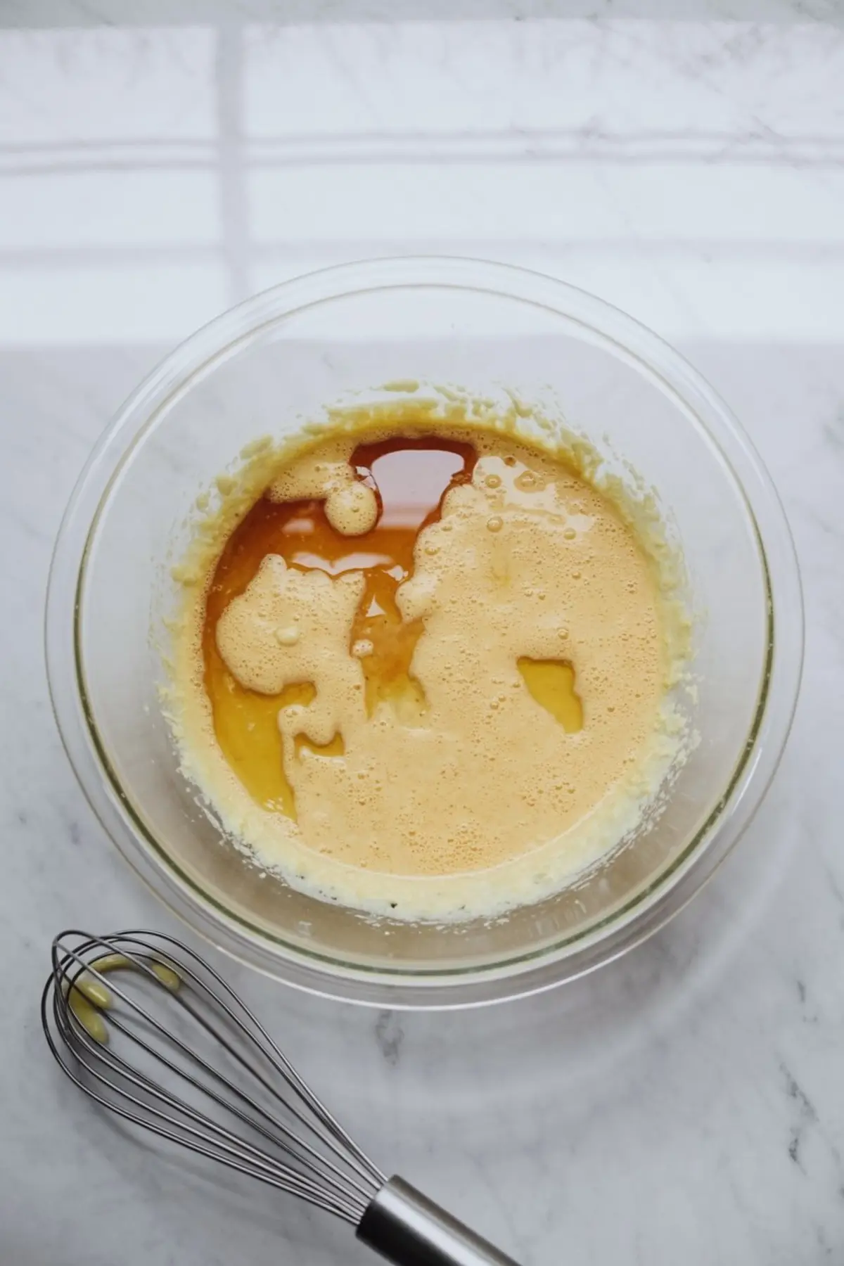 Bowl of creamed butter mixture with vanilla extract and eggs sitting on top, ready to be mixed with a metal whisk nearby.