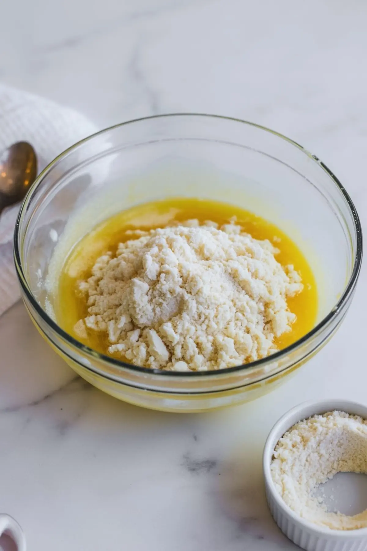 Glass mixing bowl filled with melted butter and a mound of coconut flour, placed on a white countertop beside a small dish of flour.