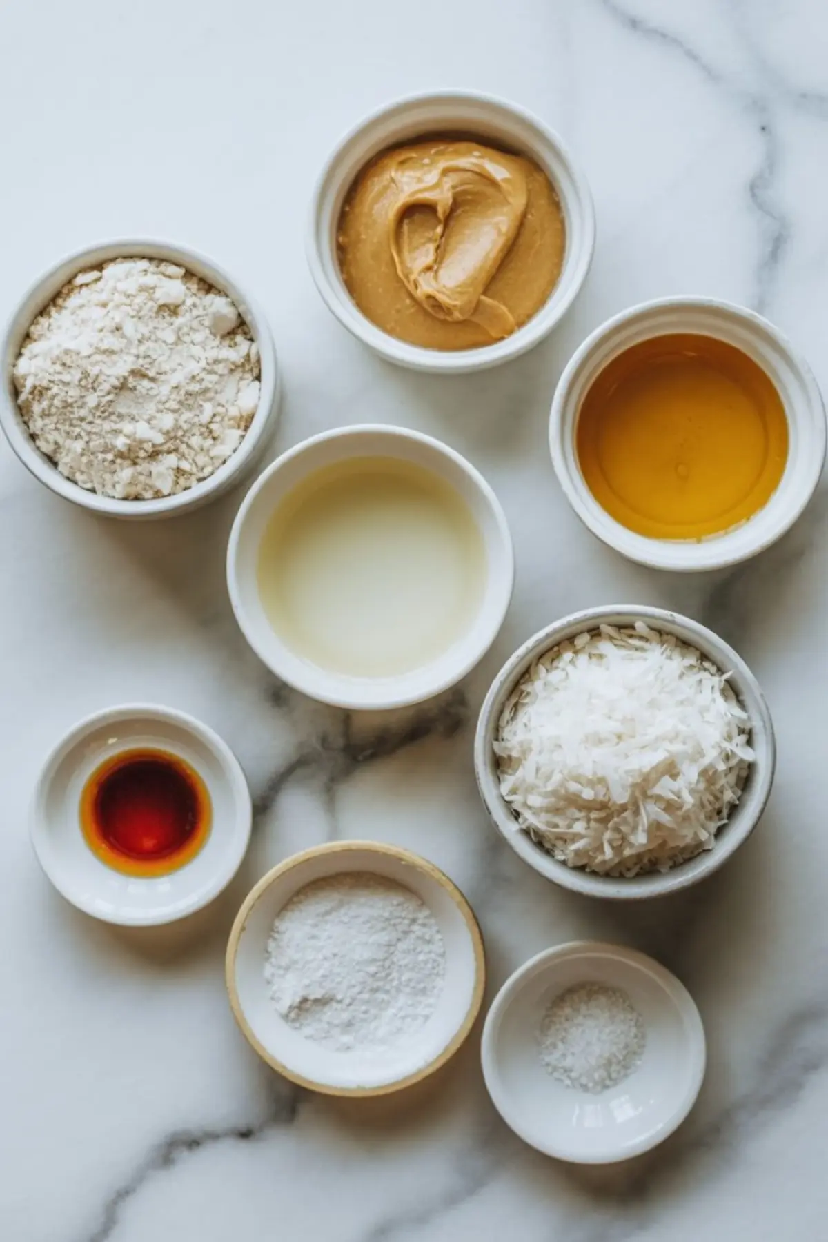 Overhead image of protein coconut bar ingredients in small bowls on a marble surface. Includes shredded coconut, peanut butter, vanilla extract, coconut oil, oat flour, maple syrup, protein powder, and salt.