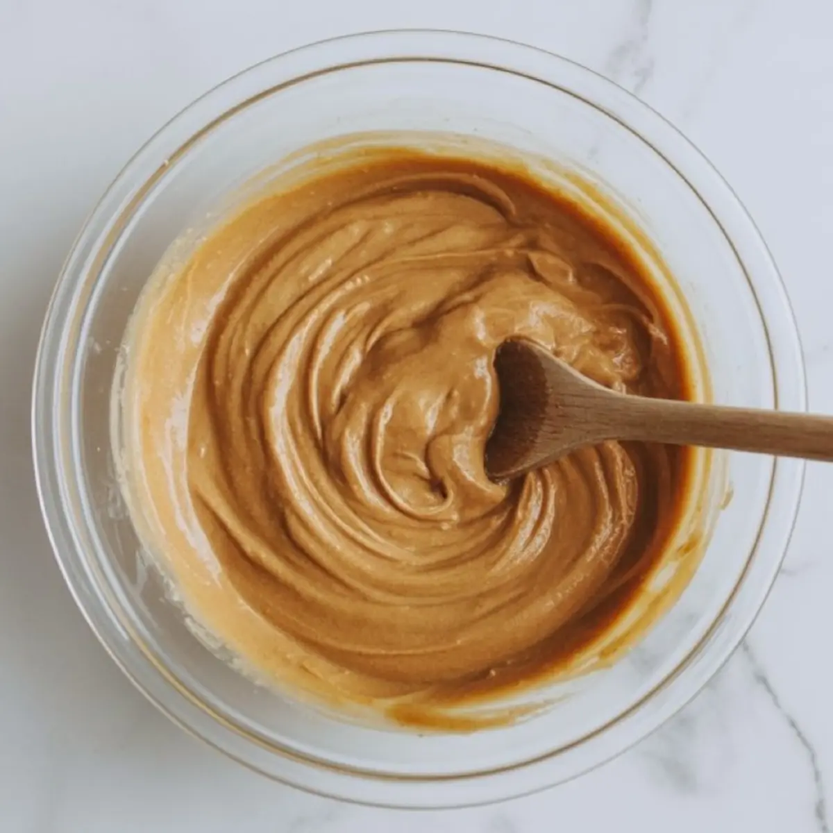 Overhead shot of thick coconut protein bar batter stirred in a glass bowl with a wooden spoon, showing creamy consistency and rich caramel-like color.