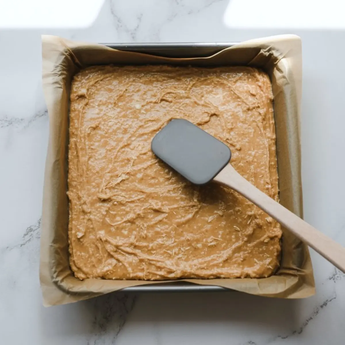 Overhead image of a baking pan lined with parchment paper filled with smoothed protein coconut bar mixture. A spatula is shown flattening the surface of the mixture before chilling or cutting.
