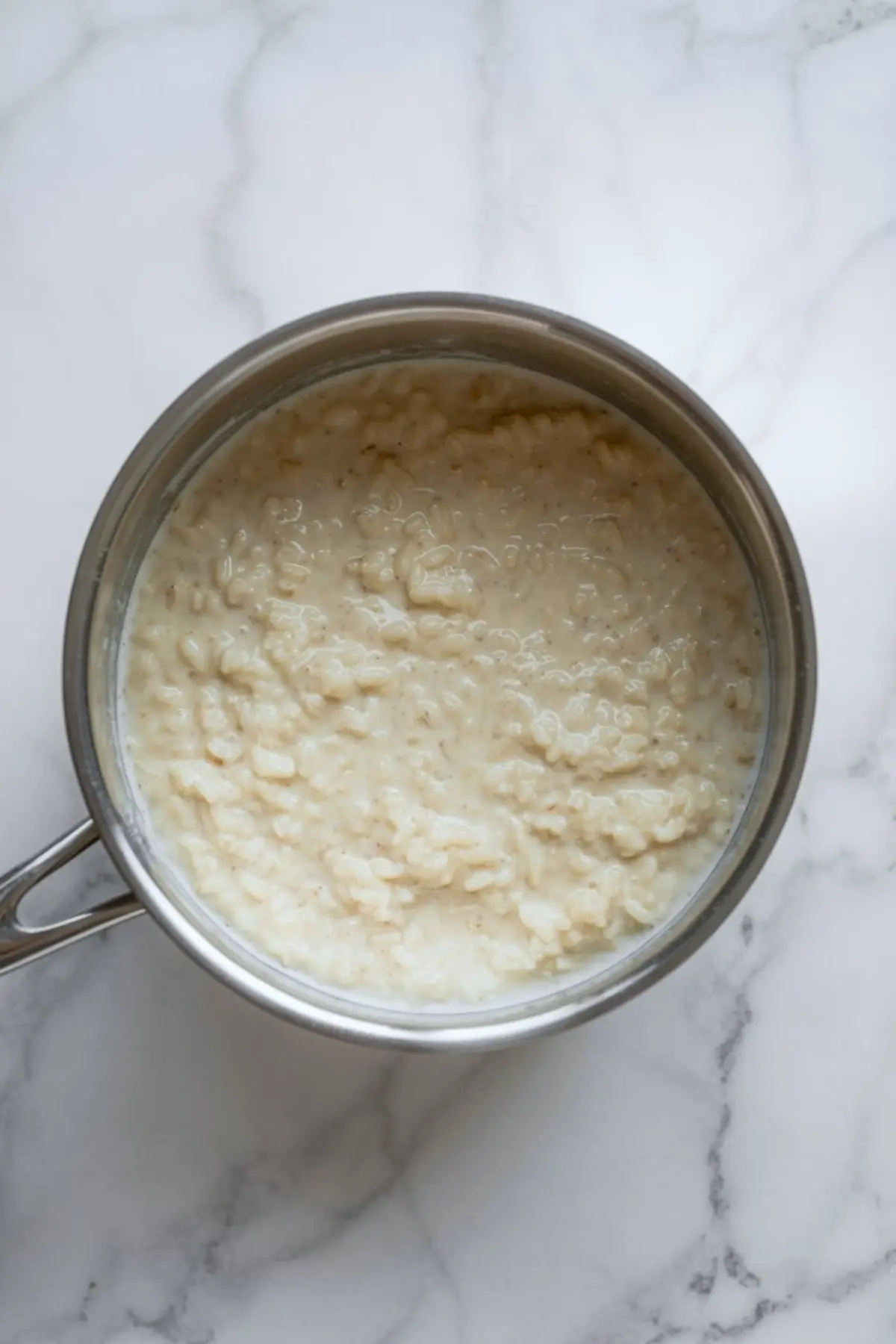 Cooked coconut rice pudding in a stainless steel saucepan on a white marble surface, showing a thick, creamy texture. The dessert appears fully cooked and ready for serving, featuring visible grains of rice mixed into a rich coconut milk base.
