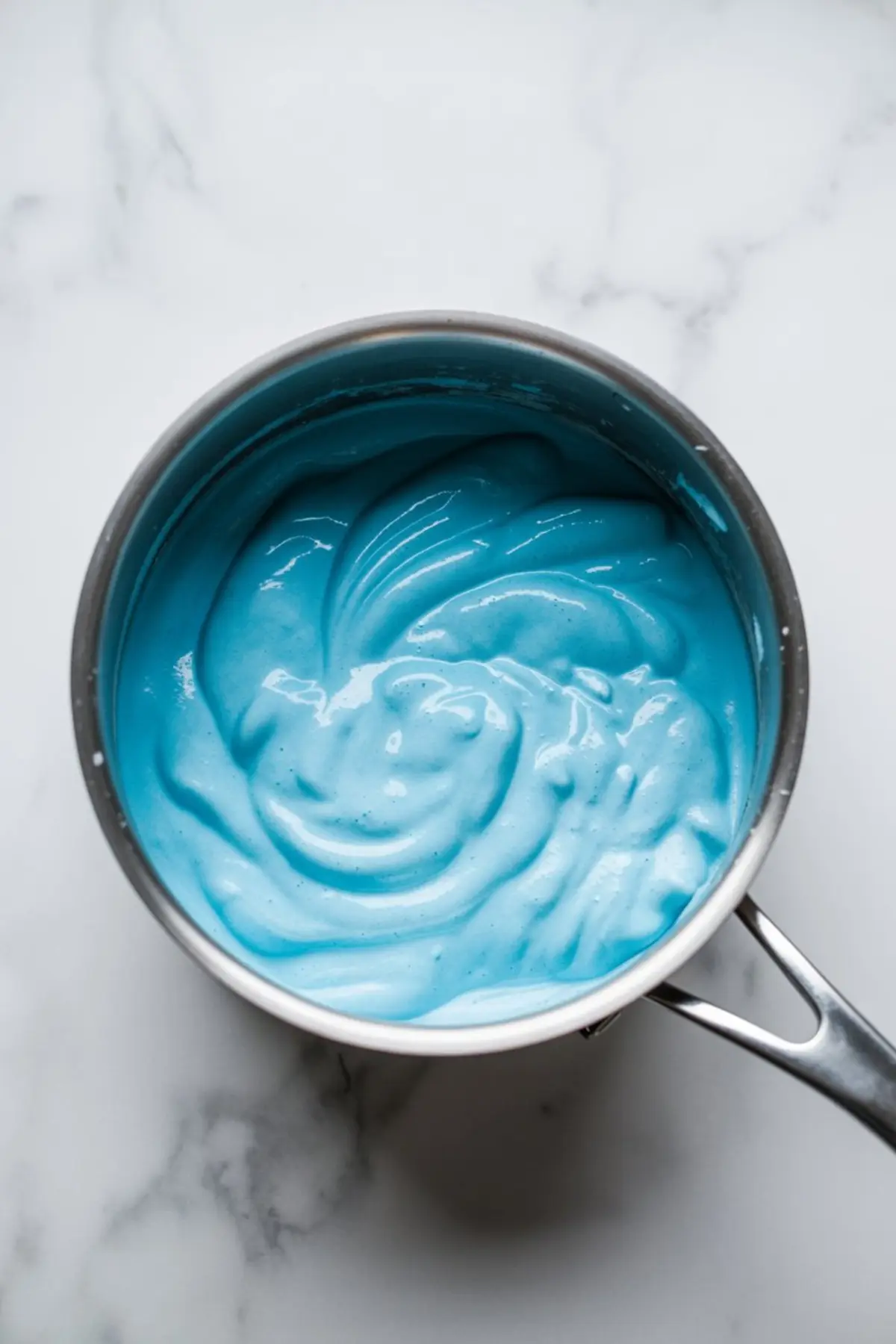Top view of a saucepan filled with smooth, bright blue melted marshmallow mixture for cotton candy rice krispie treats on a white marble background.
