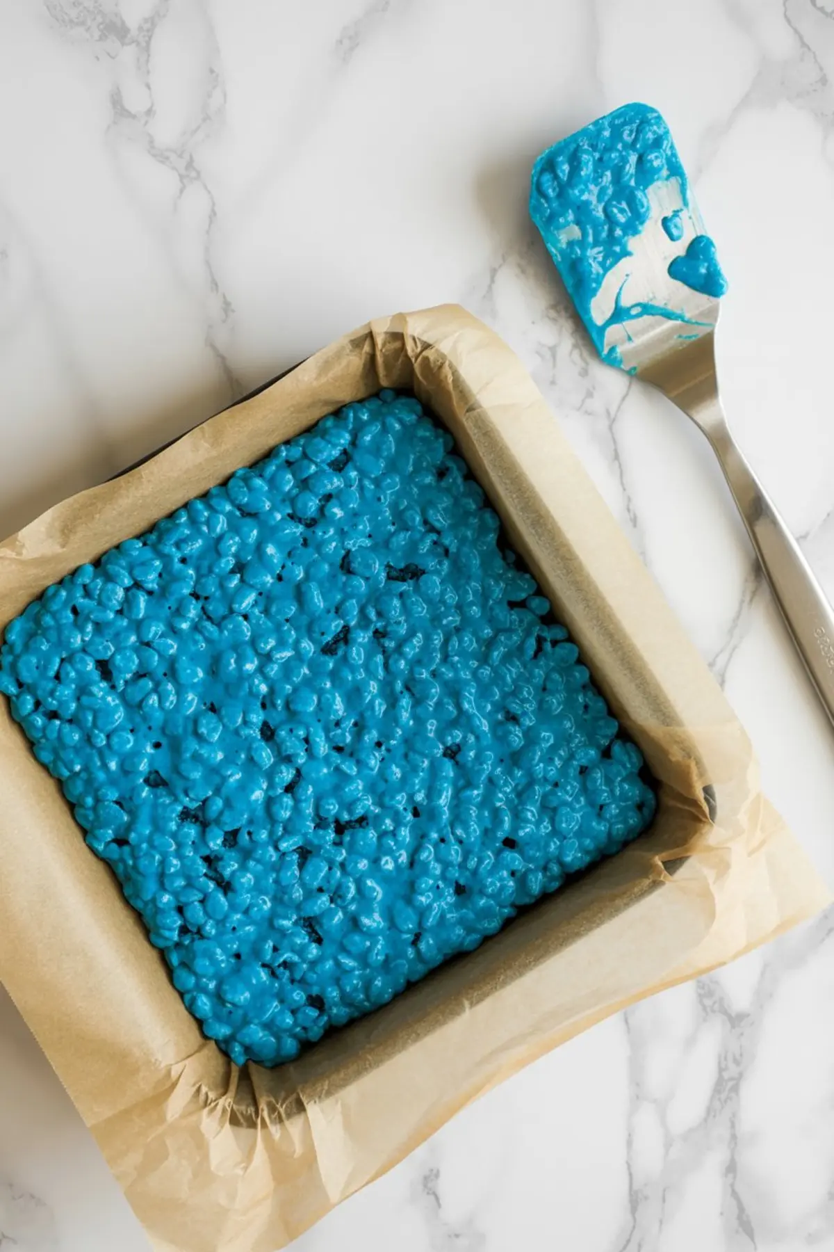 Overhead image of a baking pan lined with parchment paper and filled with vibrant blue cotton candy rice krispie mixture, next to a silicone spatula coated in the same mixture.
