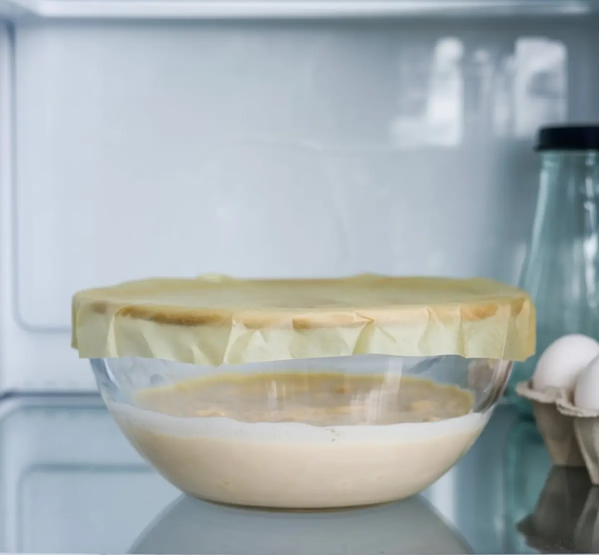 Glass mixing bowl with covered cream cheese mixture chilling in a refrigerator, next to eggs and a glass bottle.