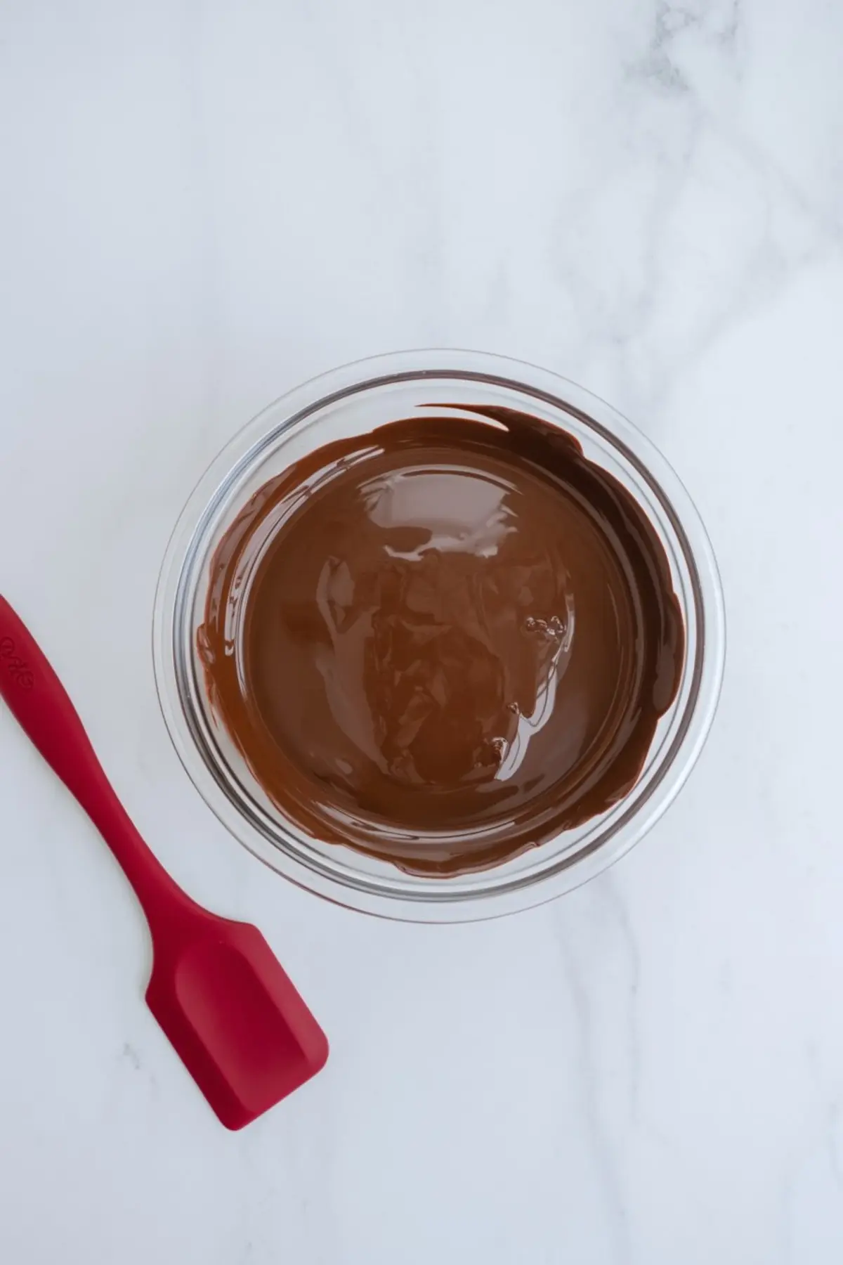 Glass bowl filled with melted dark chocolate on a white marble surface, smooth glossy chocolate texture, red silicone spatula placed next to the bowl, chocolate melting step for homemade dessert recipes.
