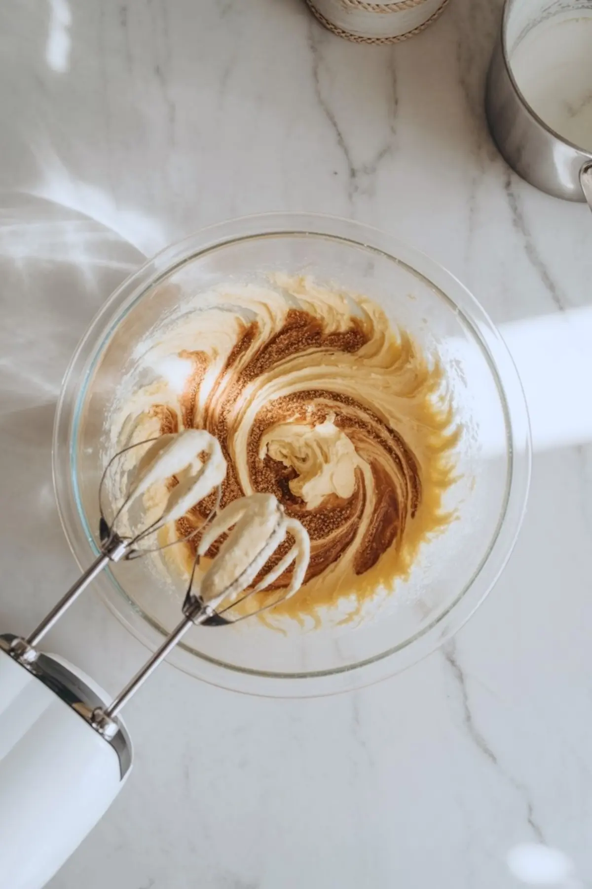 Top view of an electric hand mixer with beaters in a glass bowl of creamed butter and sugar on a marble countertop, with sunlight casting soft shadows.
