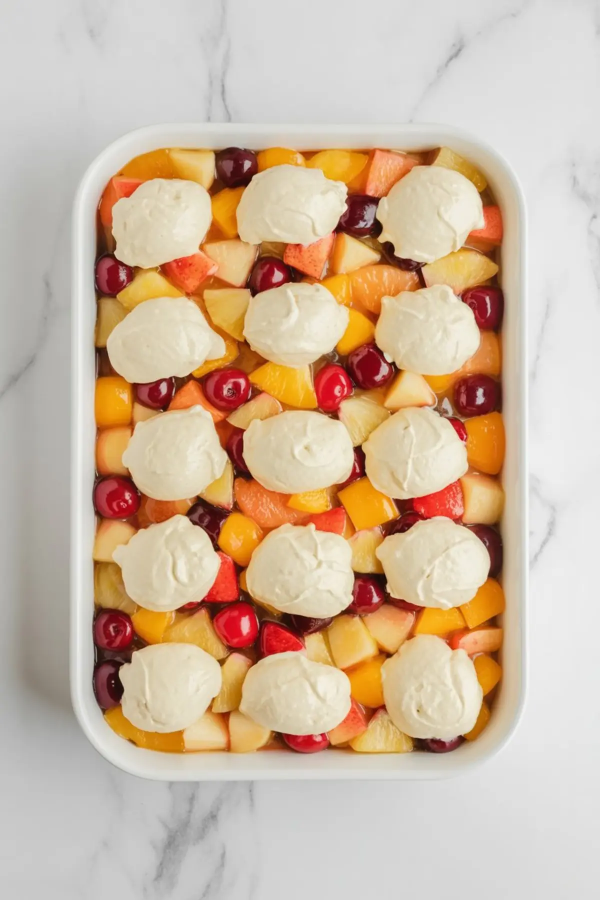 Overhead view of a white baking dish filled with fruit cocktail dessert, showing diced peaches, pears, cherries, and pineapple in syrup topped with evenly spaced dollops of creamy white topping on a marble surface.
