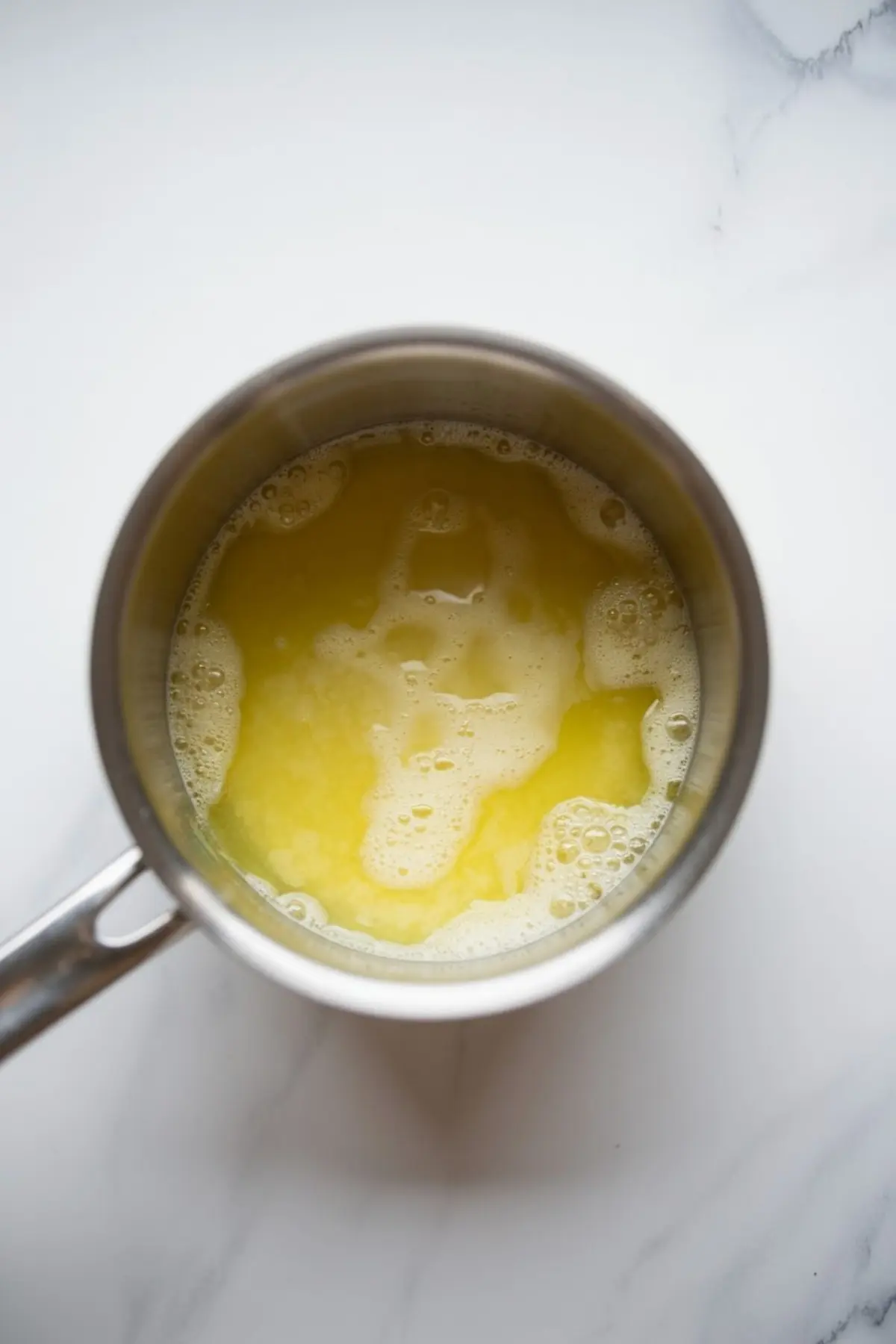 Top-down view of melted butter foaming in a small metal saucepan, showing glossy yellow butter with bubbles forming on the surface on a white marble countertop.
