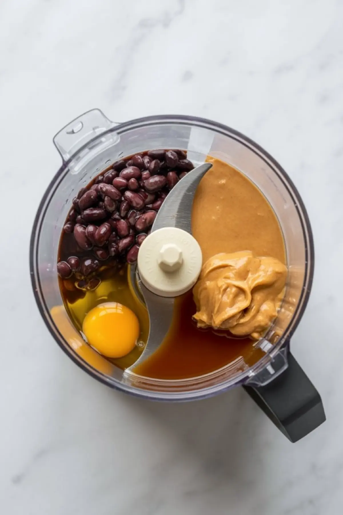 A food processor bowl contains black beans, peanut butter, egg, vanilla extract, and oil arranged before blending for black bean brownie batter.
