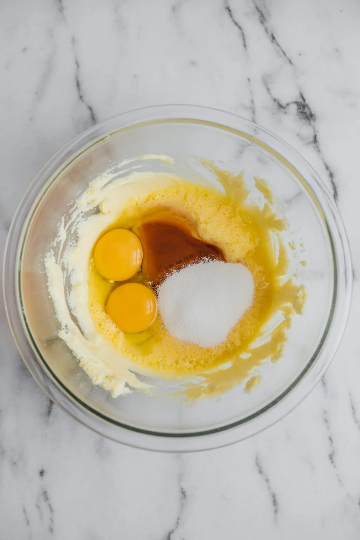 Overhead view of a glass mixing bowl containing eggs, granulated sugar, and vanilla extract on top of a whipped butter base, ready to be mixed for hazelnut cake batter.
