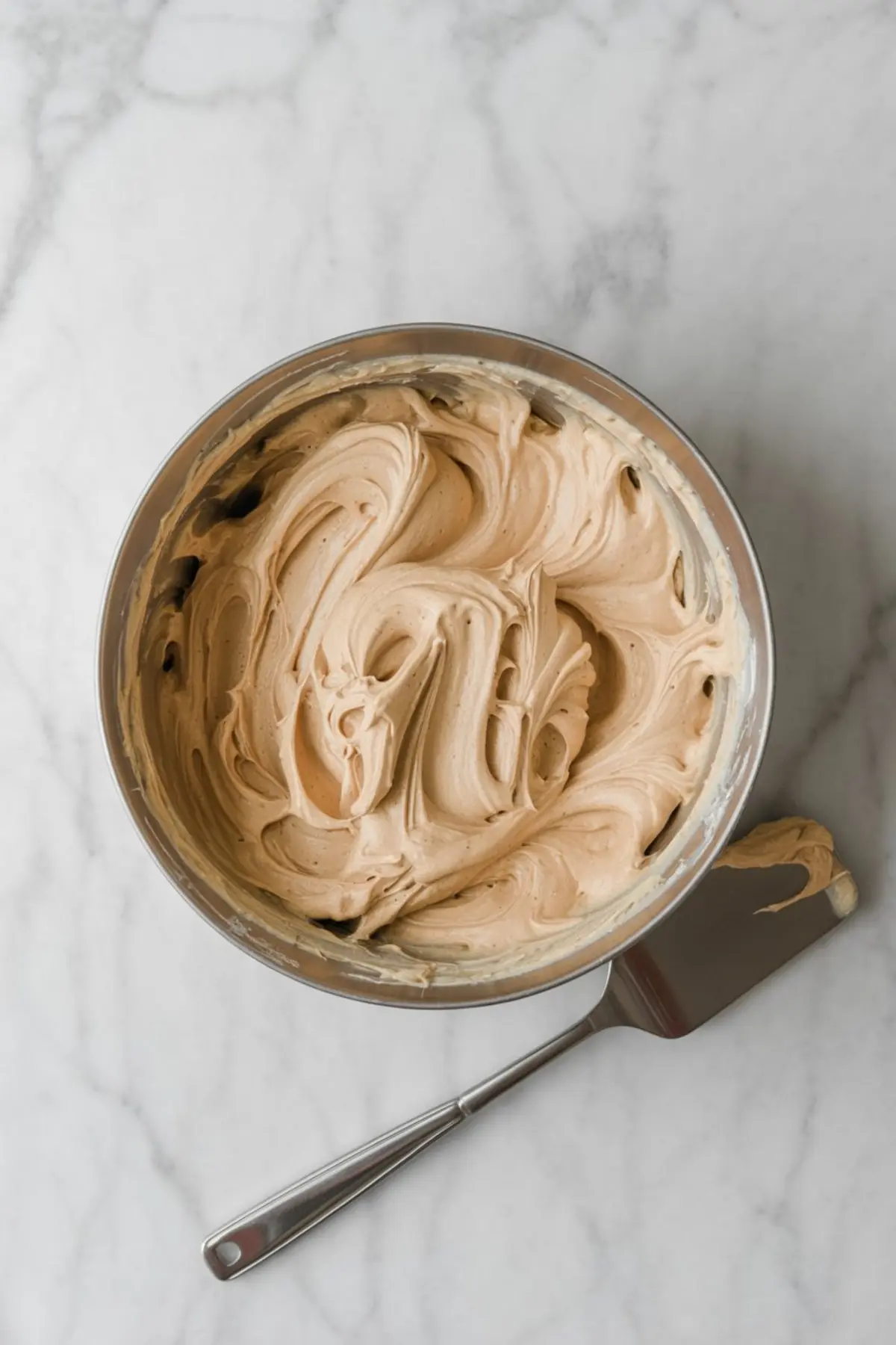 Bowl of whipped hazelnut frosting with soft, airy texture swirled throughout, placed on a marble surface next to a silver spatula with a dollop of frosting on the edge.
