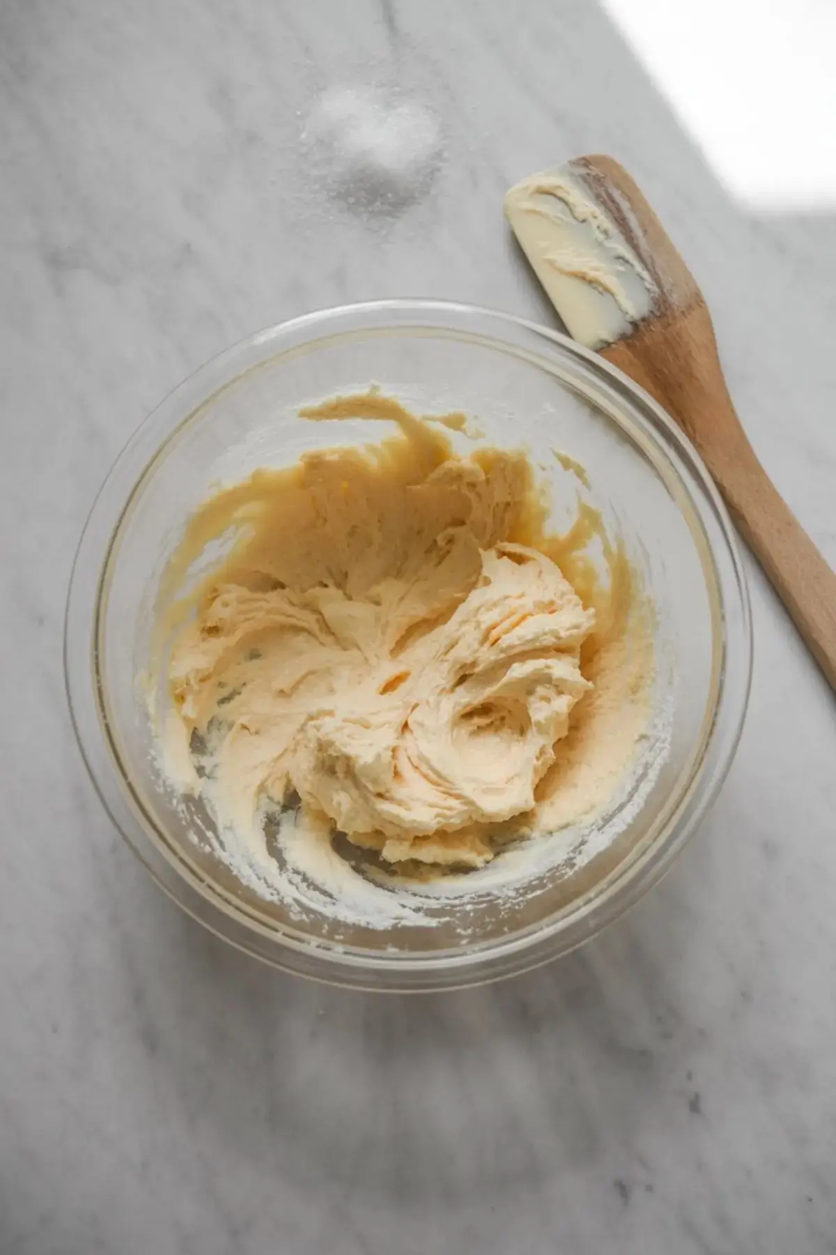 Glass bowl containing creamed butter and sugar mixture with a light fluffy texture, resting on a marble countertop beside a wooden spatula with remaining mixture.
