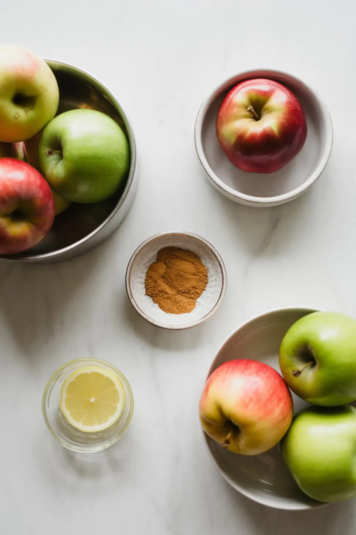 Fresh red and green apples arranged in ceramic bowls beside a glass of lemon water and a small dish of ground cinnamon on a white countertop, showing the essential ingredients for baked apple chips.
