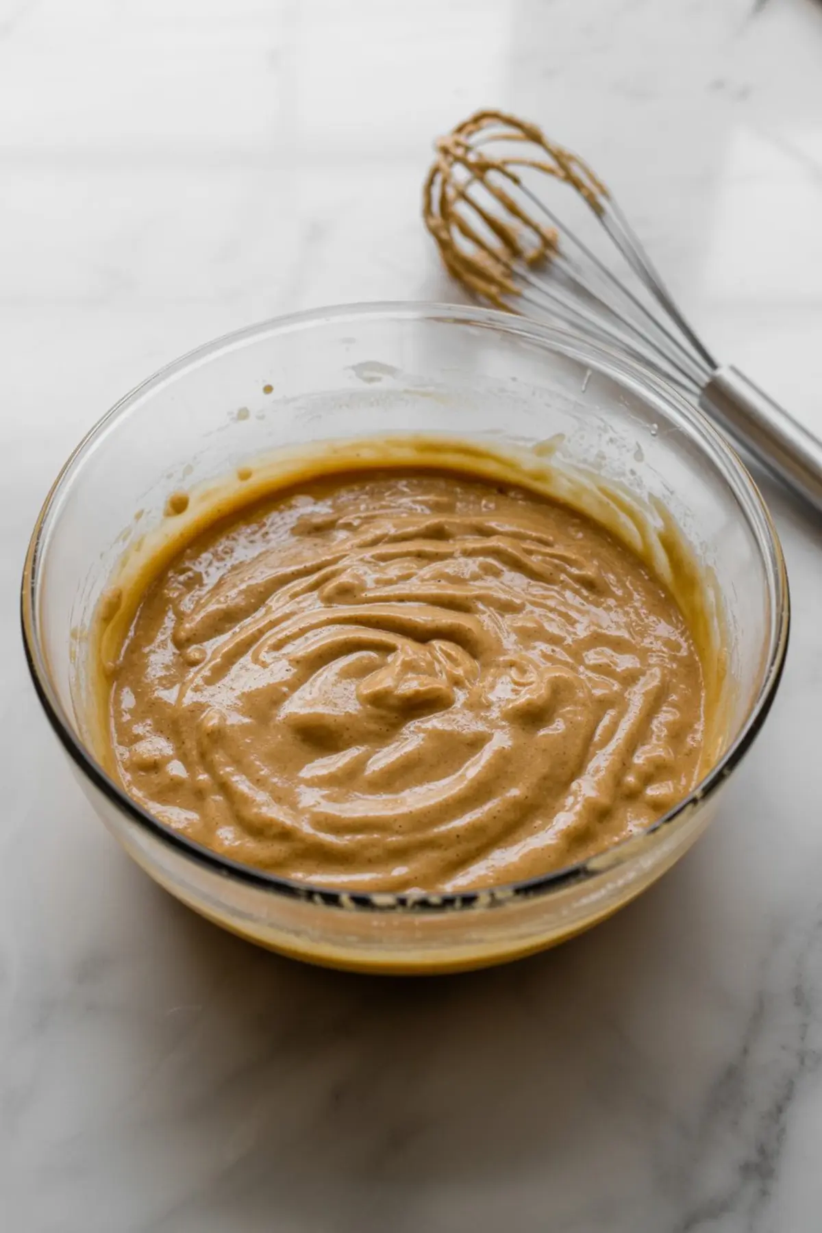 Glass mixing bowl filled with light brown muffin batter resting on a marble countertop beside a whisk coated in batter.
