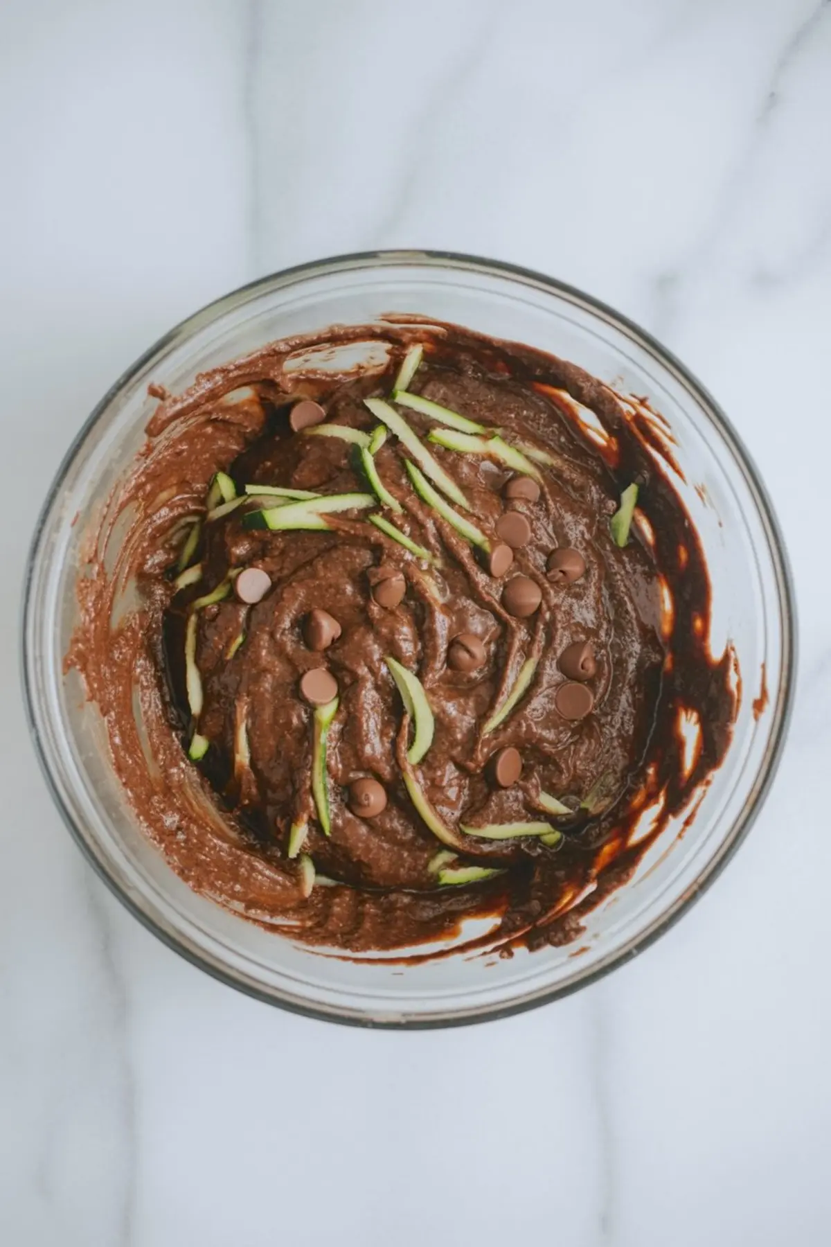 Top view of chocolate zucchini muffin batter in a glass bowl, featuring shredded zucchini and semi-sweet chocolate chips swirled throughout.
