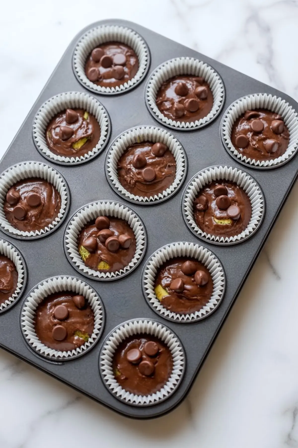 Muffin tin filled with raw chocolate zucchini muffin batter in white paper liners, each topped with chocolate chips, ready for baking.
