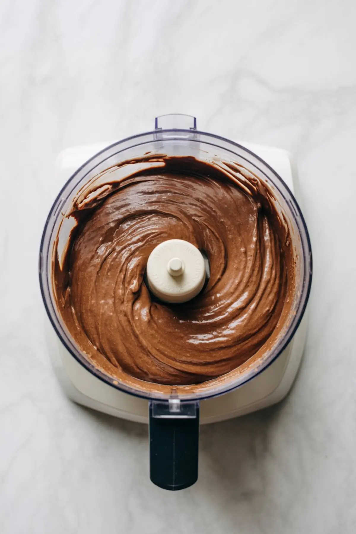 Chocolate chickpea cookie dough blended in a food processor, showing smooth cocoa banana batter prepared for healthy high protein cookies on a light marble countertop.
