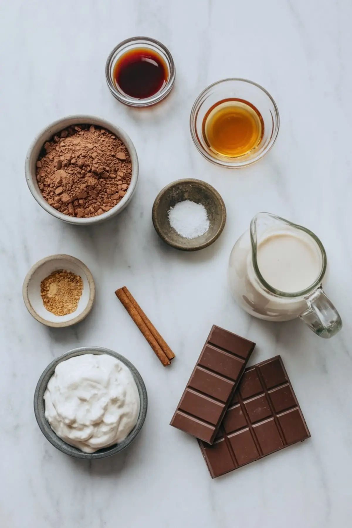 Flat lay of healthy hot chocolate ingredients on a marble surface, including cocoa powder, dark chocolate bars, coconut cream, cinnamon sticks, sea salt, maple syrup, vanilla extract, coconut milk, and coconut sugar.