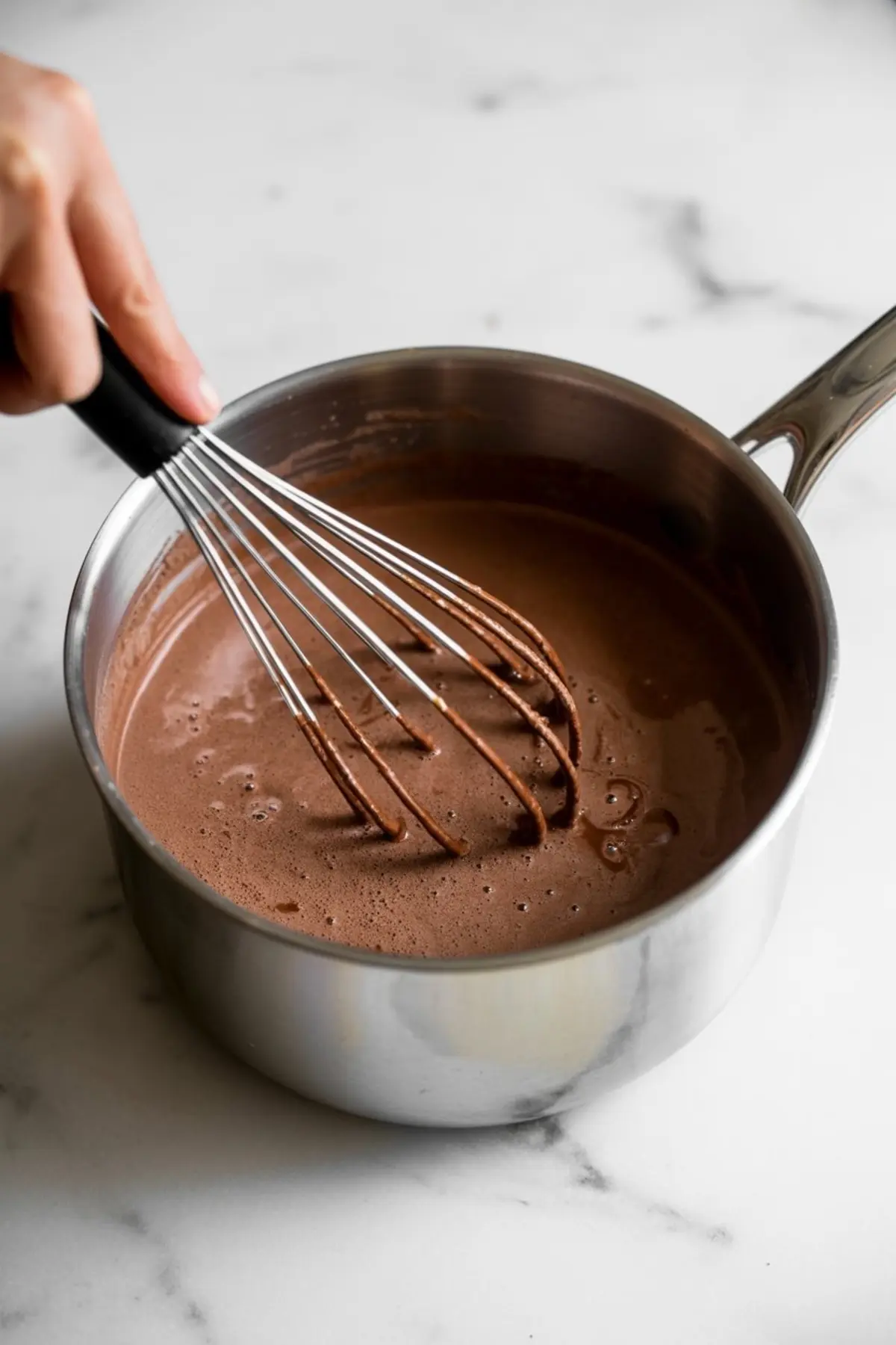 Whisking rich hot chocolate in a stainless steel saucepan, blending cocoa and plant milk into a smooth, frothy drink on a marble surface.