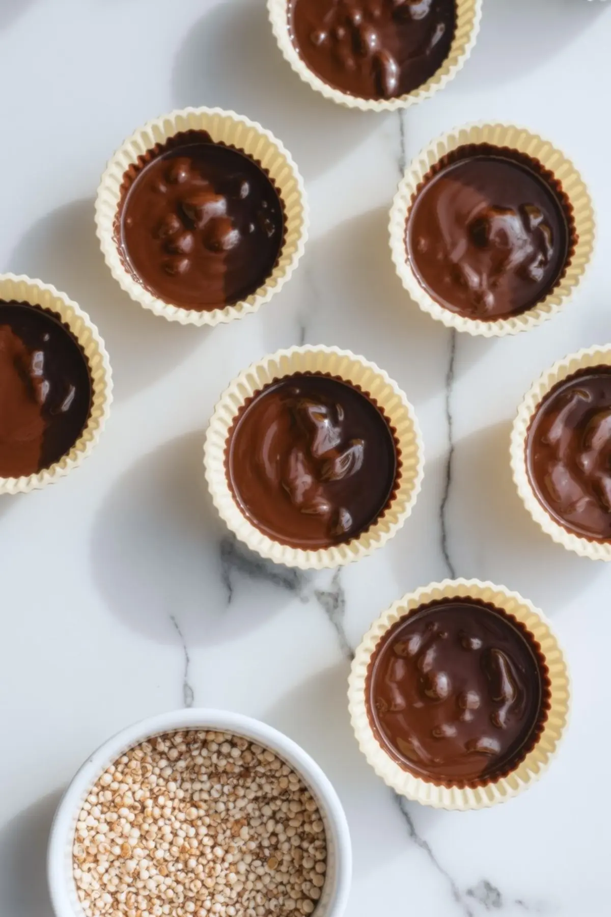 Silicone baking cups filled with melted chocolate and puffed quinoa mixture on a marble surface, prepared for healthy peanut butter cups.