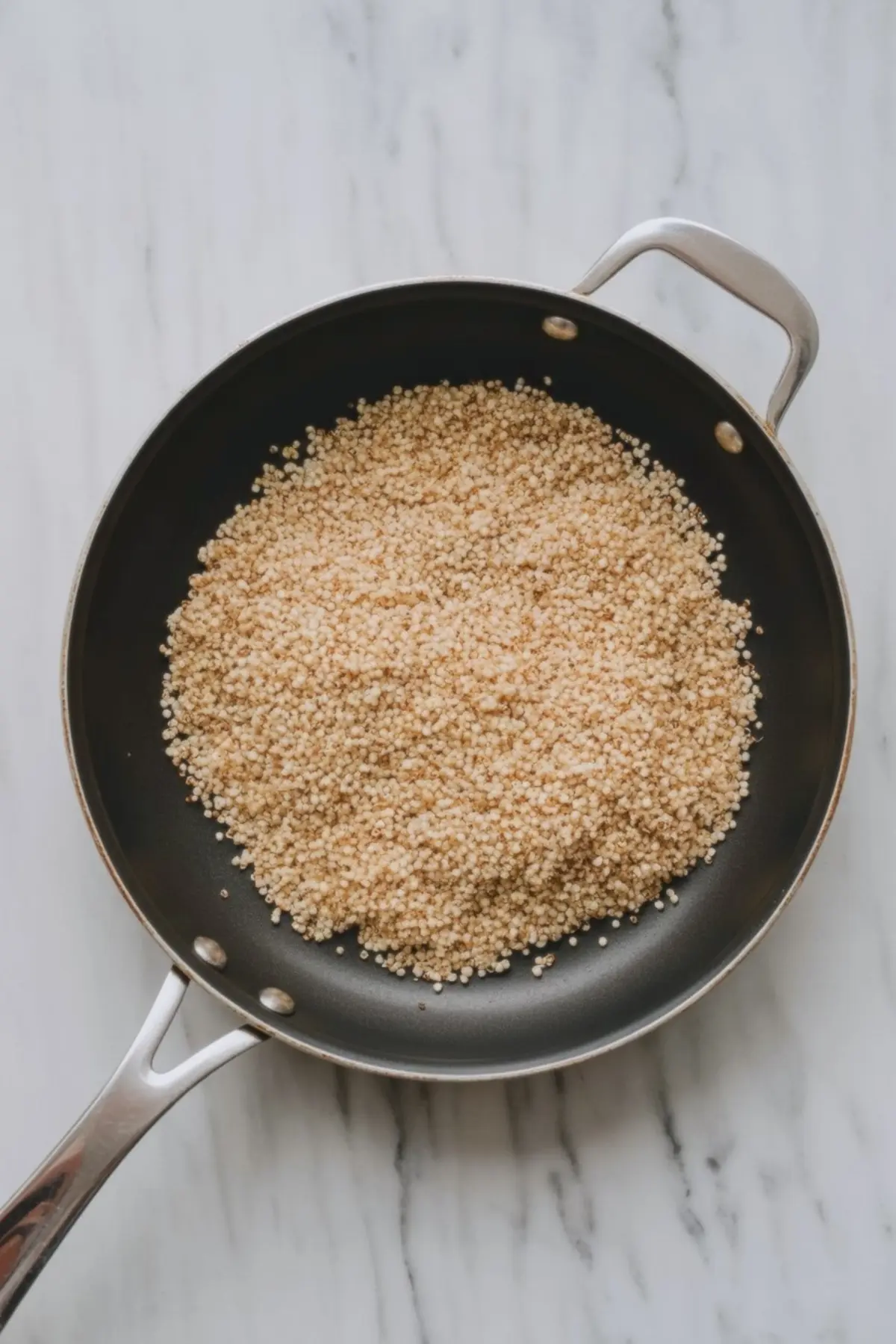 Non-stick pan filled with golden toasted puffed quinoa on a white marble background, ready for chocolate quinoa treats.