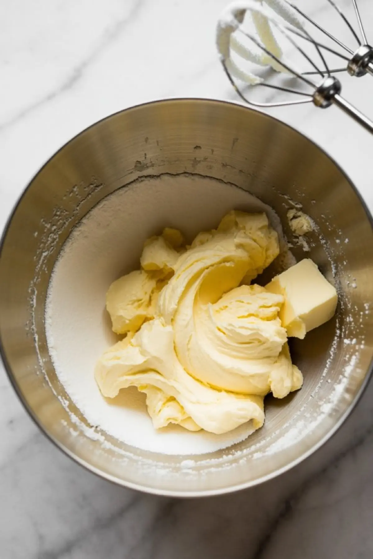 Close-up of creamed butter and sugar mixture in a metal mixing bowl with a hand mixer in the background, on a white marble countertop.
