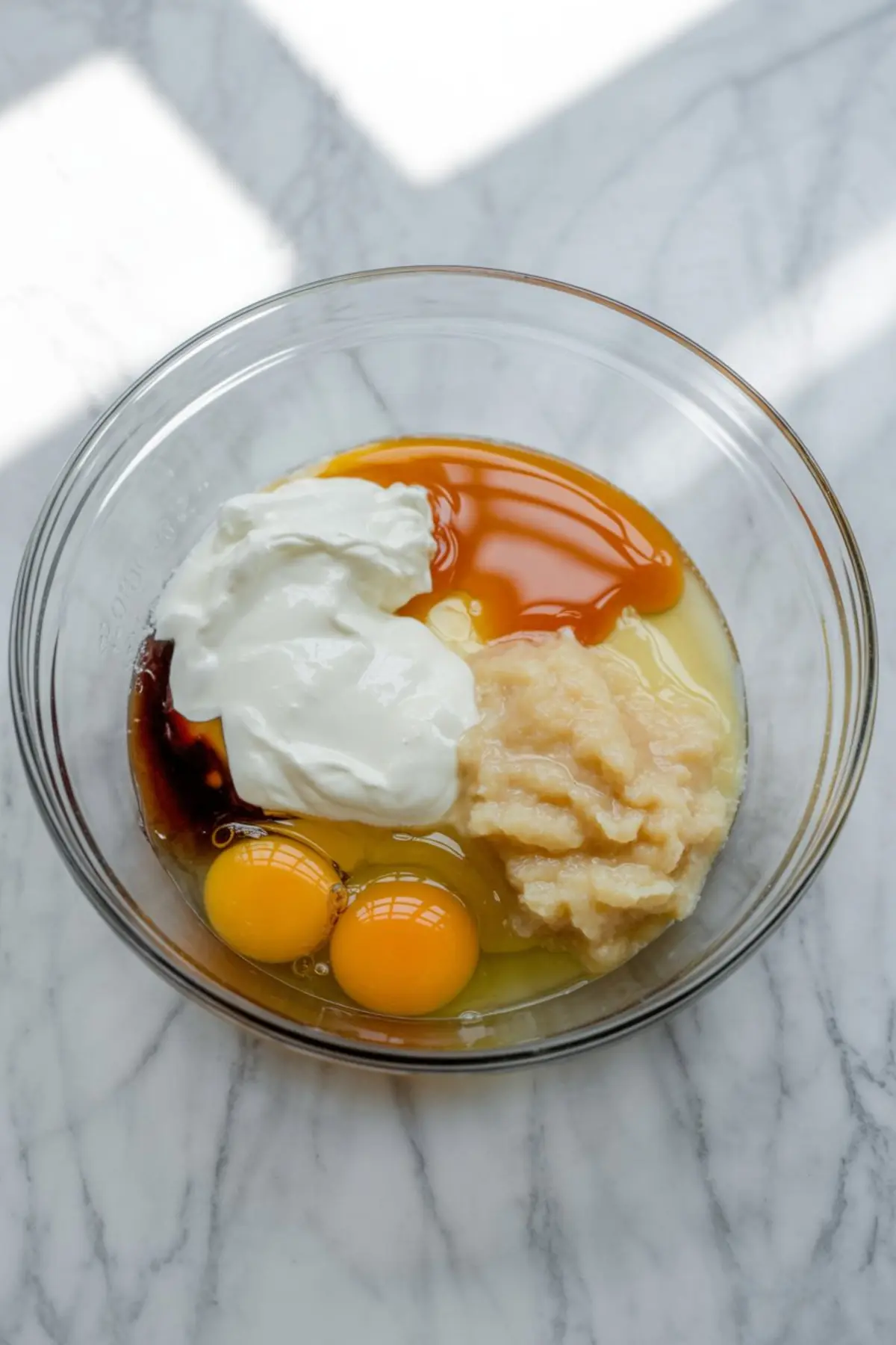 Wet ingredients rest in a glass mixing bowl, including eggs, Greek yogurt, applesauce, vanilla extract, oil, and honey, shown before mixing on a bright marble background.
