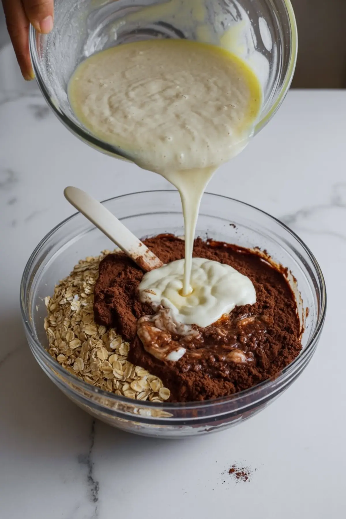 Creamy wet batter pours from a glass bowl into a larger bowl of dry ingredients with oats and cocoa powder, showing the mixing step for protein brownie batter on a marble countertop.
