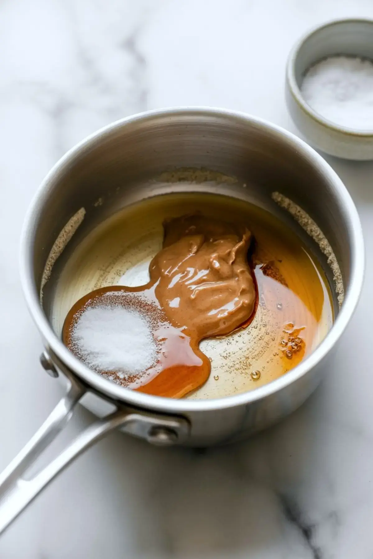 Stainless steel saucepan on a marble countertop filled with creamy peanut butter, maple syrup, and a scoop of baking soda before mixing for a protein bar recipe.
