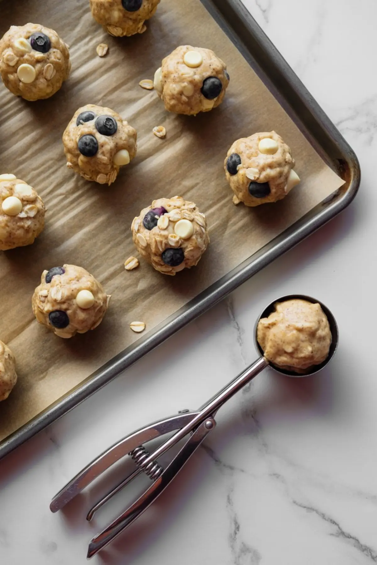 Scooped cookie dough balls on a parchment-lined baking tray, featuring blueberries, oats, and white chocolate chips. A cookie scoop rests on the marble countertop, showcasing healthy breakfast cookie prep before baking.