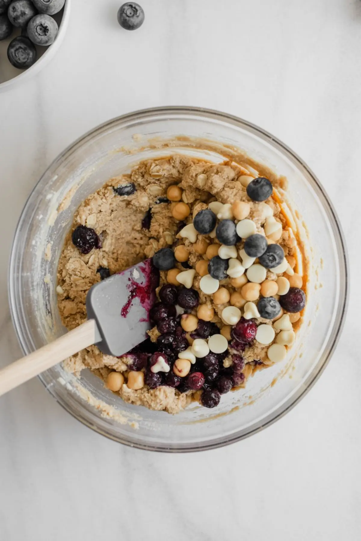 Large mixing bowl with a partially mixed cookie dough containing blueberries, chickpeas, and white chocolate chips. A silicone spatula rests in the bowl, capturing the vibrant and textured ingredients for a nutrient-dense cookie recipe.