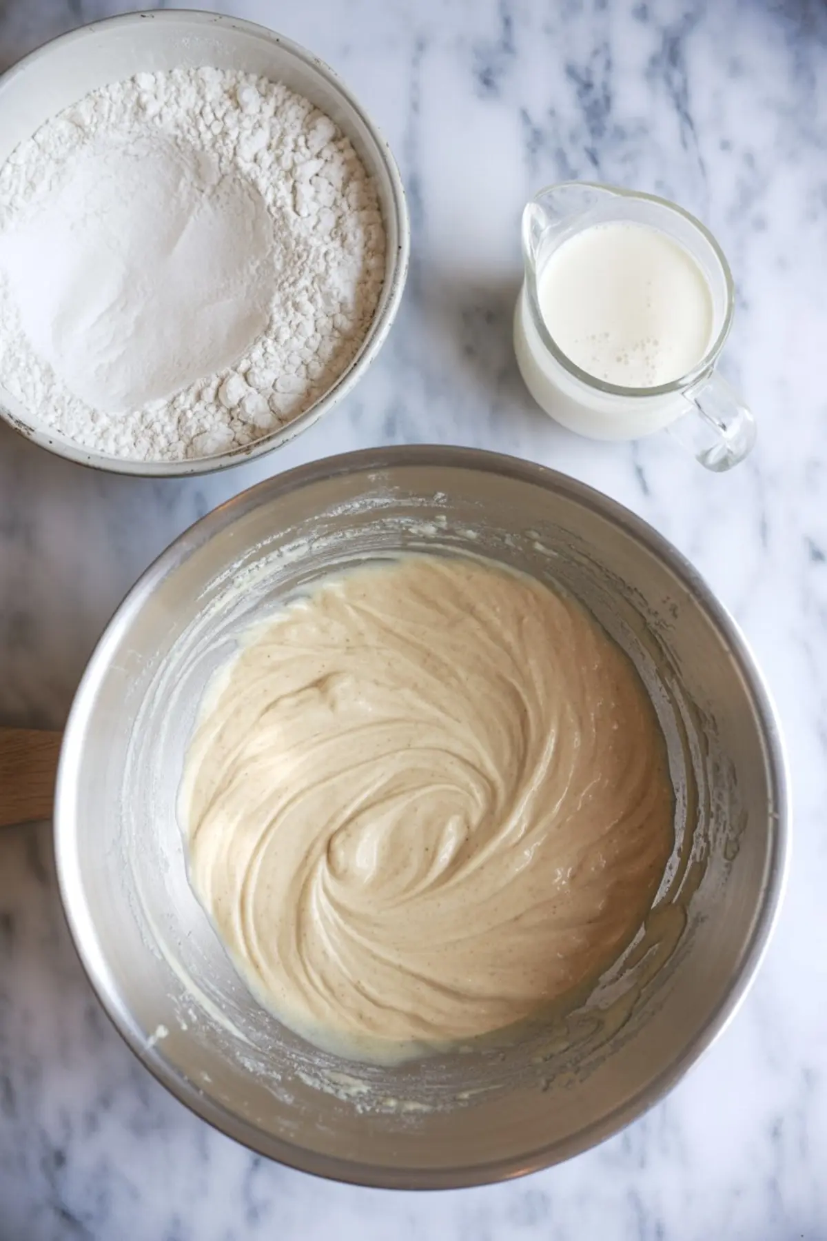Mixed lemon cake batter in a metal bowl with visible vanilla specks, set beside bowls of flour and buttermilk on a marble countertop.
