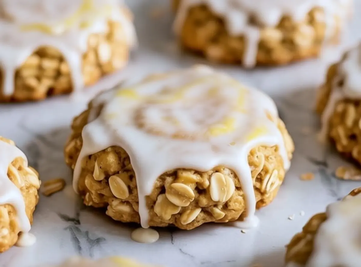Close-up of a thick lemon oatmeal cookie topped with glossy white icing, showcasing visible oats and a soft, chewy texture on a marble surface.
